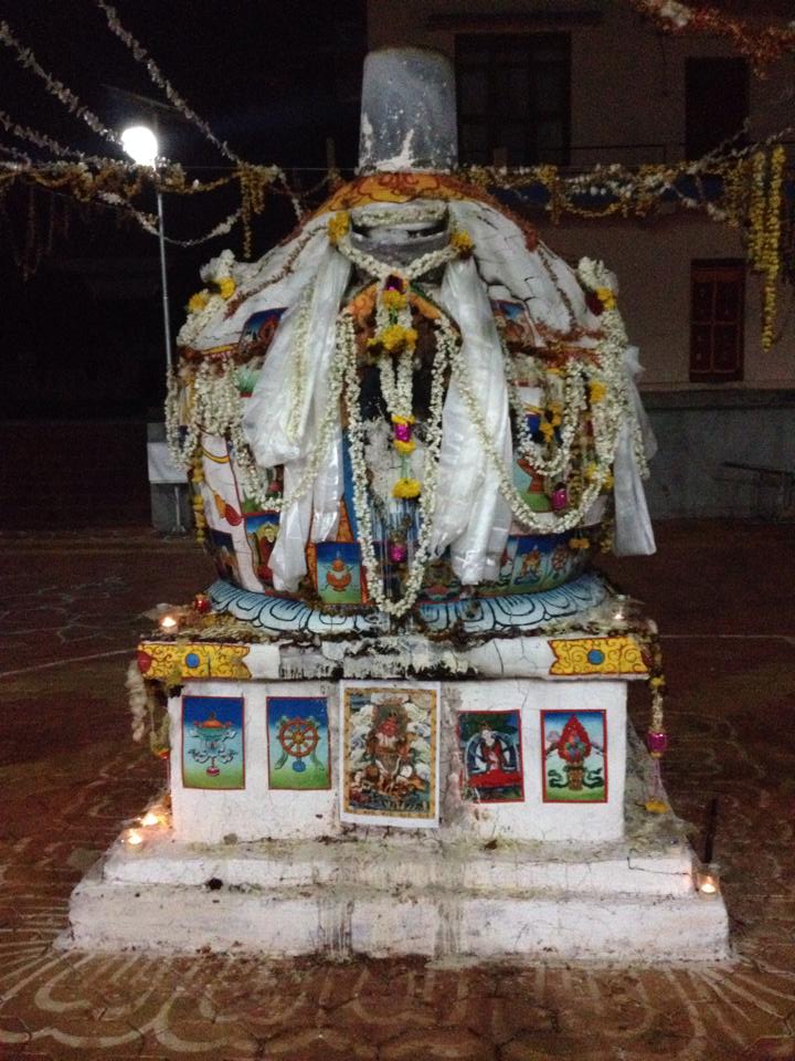 A whitewashed stupa at night, draped in white khatas and marigold garlands, with butter lamps at its base